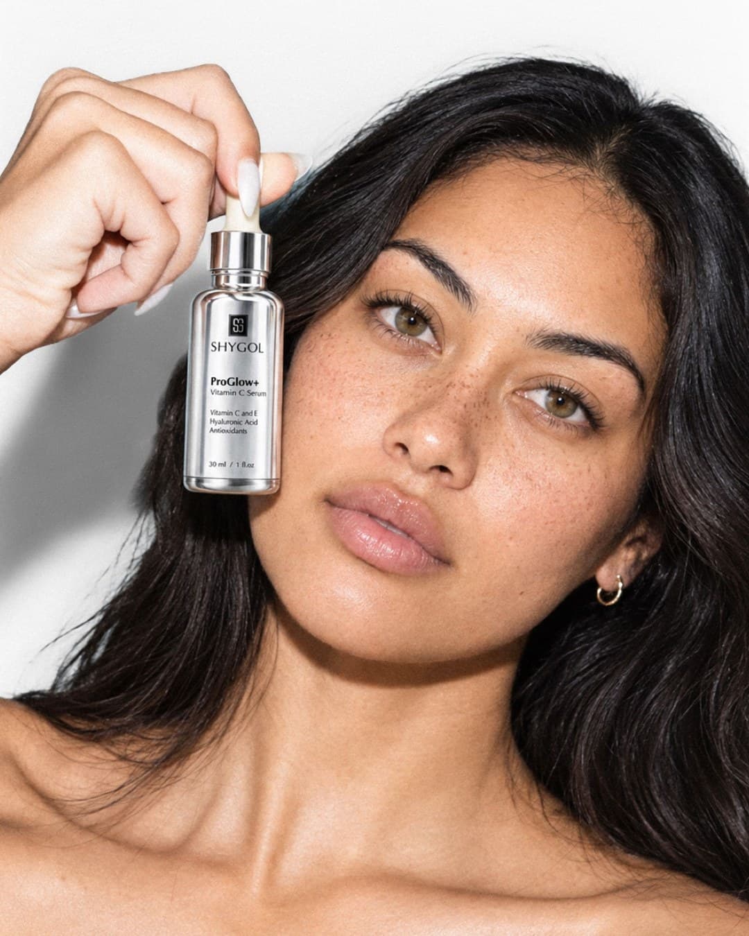 Shygol model shot: freckled model holding a ProGlow+ serum bottle up beside her cheek and looking at the camera
