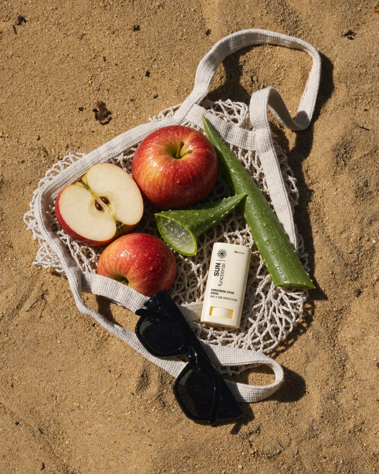 Overhead flat lay of a SUN functional sunscreen stick, apples and aloe leaves spilling out of a mesh beach bag on sand